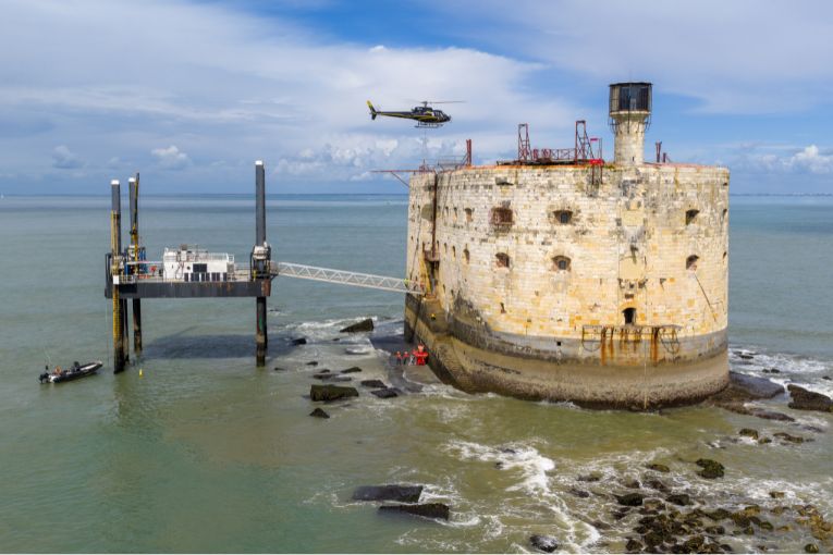 Depuis les airs, un hélicoptère transporte des bennes contenant du béton coulé fourni par Edycem, en immergé directement sous l’eau. [©Imagine Créations - Groupe ETPO - Architecture Patrimoine – BRL Ingénierie]